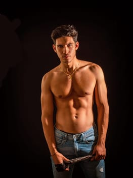 Handsome muscular shirtless young man standing confident, looking at camera on black background