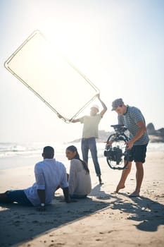 Bounce those sun rays. a man holding up a sun screen to dim the sun rays on two models that are being filmed by a videographer