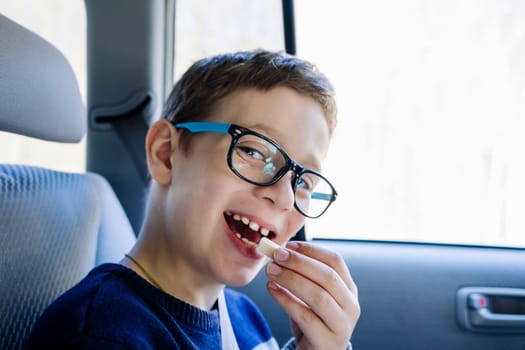 A happy Caucasian boy of school age rides in the back seat of a car and eats candy. A schoolboy with glasses. The child is wearing seat belts, and he is traveling with his family by car