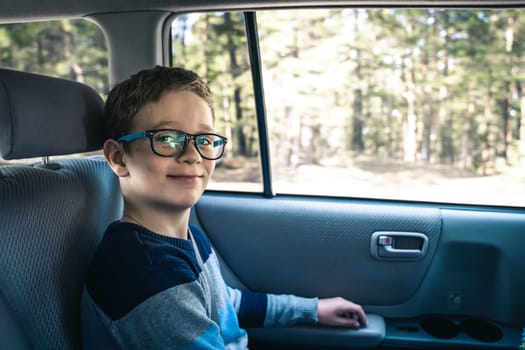 A happy Caucasian boy of school age rides in the back seat of a car and looks at the camera . A schoolboy with glasses. The child is wearing seat belts, and he is traveling with his family by car