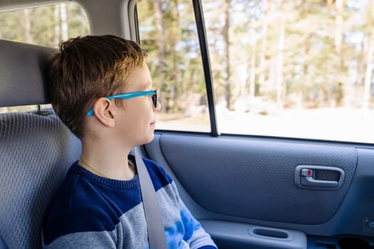 a Caucasian boy of school age rides in the back seat of a car and looks out the window. The child is wearing seat belts and he is traveling with his family by car