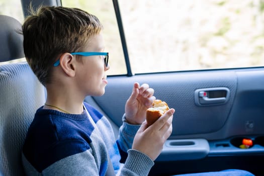 A happy Caucasian boy of school age rides in the back seat of a car and eats a sandwich. A schoolboy with glasses. The child is wearing seat belts, and he is traveling with his family by car