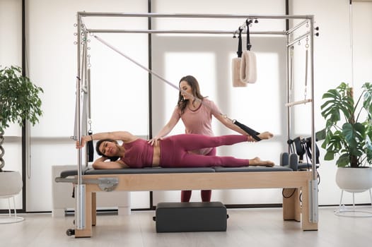 Asian woman doing pilates with trainer on cadillac reformer