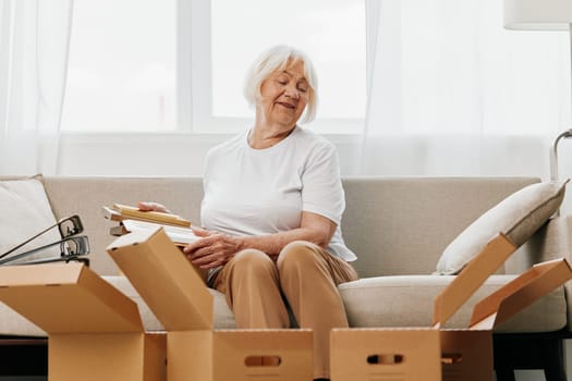 elderly woman sits on a sofa at home with boxes. collecting things with memories albums with photos and photo frames moving to a new place cleaning things and a happy smile. Lifestyle retirement. High quality photo