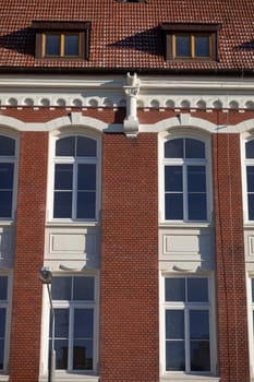 A section of the curtain wall of a historic red brick building with white inserts around the windows. Two roof dormers.