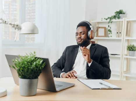 An African-American man sits at his desk in front of his laptop, wearing headphones and chatting on a video call, listening to music. The concept of student business training and online work. High quality photo