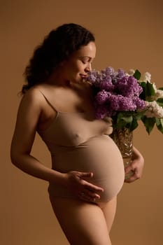 Attractive pregnant woman in beige underwear, posing with beautiful bunch of lilacs, isolated on beige background. Fashion portrait of authentic young gravid woman expecting a baby. Pregnancy 30 weeks