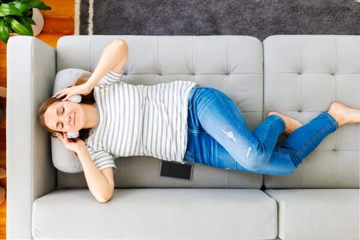 Peaceful woman in modern wireless headphones laying, relaxing on a comfortable couch, listening to music. Happy calm woman wearing headphones relaxing on a comfortable sofa, enjoy quality sound