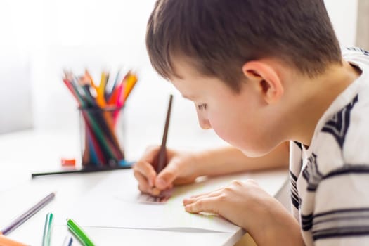 A child boy draws on white paper with colored pencils while sitting at a table.