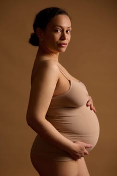 Side portrait of a beautiful Latina pregnant woman, expectant young mother in beige lingerie, holding hands on her belly, looking confidently at camera, isolated studio background. Pregnancy 30 week
