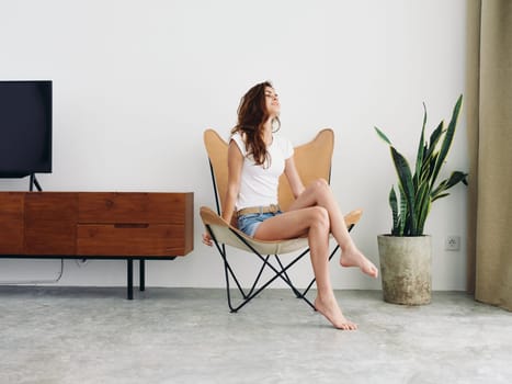 Woman sitting in leather armchair smile with teeth Lifestyle in white T-shirt and blue denim shorts, resting at home stylish modern interior with white walls, copy space. High quality photo