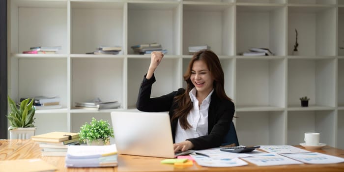 Happy excited successful businesswoman triumphing in office. Young asian got good work result, business woman is happy and celebrating victory holding hands up looking and reading from laptop screen.