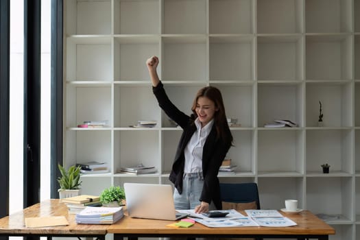 Happy excited successful businesswoman triumphing in office. Young asian got good work result, business woman is happy and celebrating victory holding hands up looking and reading from laptop screen.