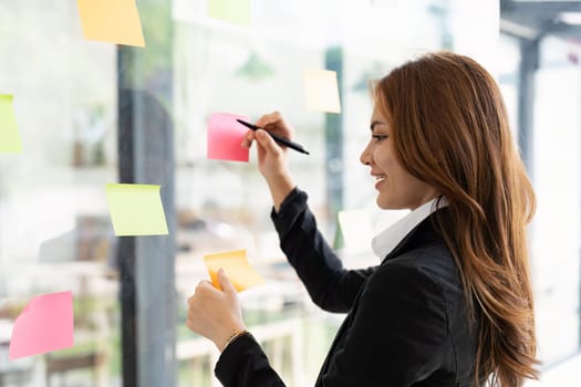 Asian businesswoman creating project plan on office wall with paper notes. Financial and Marketing projects.