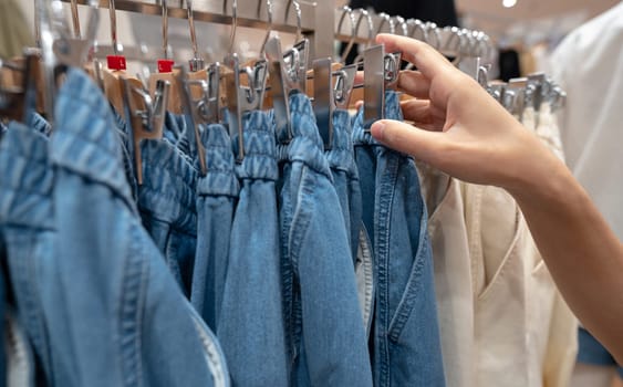 Woman shopping denim pants in clothing store. Woman choosing clothes. Jeans on hanger hanging on rack in clothing store. Fashion retail shop inside shopping mall. Clothes on hangers in a clothes shop.