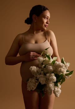 Confident gravid woman in beige underwear, holding hand on her pregnant belly, looking aside, posing with a bunch of while blooming lilacs, isolated on beige background. Pregnancy Maternity Gynecology