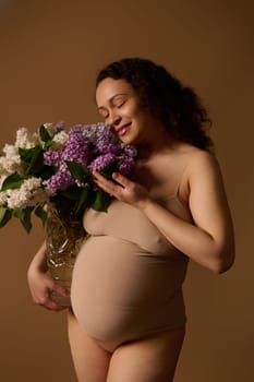 Delightful gravid woman mother in beige lingerie, posing with purple and while blooming lilacs, isolated on beige background. Pregnancy. Maternity. Gynecology. Women's health and fertility concept