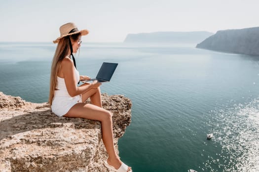 Successful business woman in yellow hat working on laptop by the sea. Pretty lady typing on computer at summer day outdoors. Freelance, travel and holidays concept.