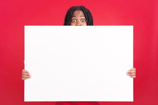 Surprised african woman hiding behind a blank panel in studio with red background