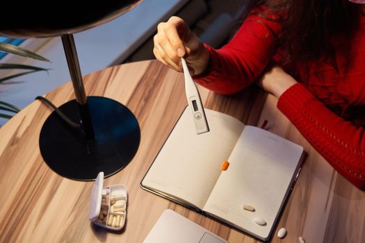 sick young woman with pills and thermometer on a table working on a laptop from home on quarantine isolation. Online work, stay home concept