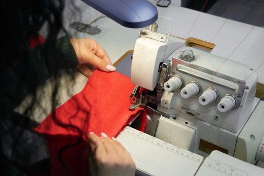 Young woman using professional overlock sewing machine in workshop. Equipment for edging, hemming or seaming clothes at tailors shop