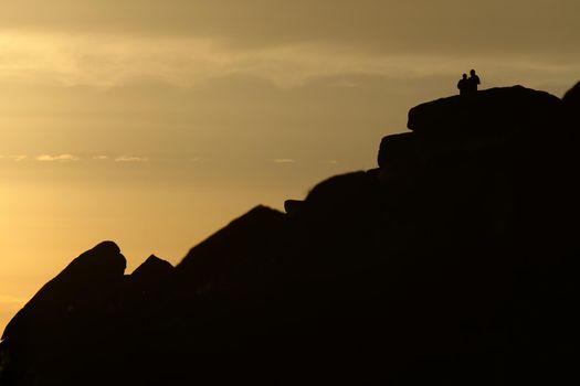 Dramatic rock formations of stanage edge in the peak district are silhouetted against the golden sky of a beautiful sunset. A couple stand at the top