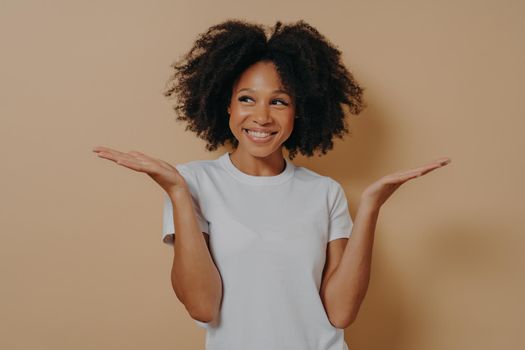 Young cheerful african woman can not make decision, spreading palms, dark skinned female with curly hair having no idea what happened while posing isolated over beige background. Hesitation and doubts