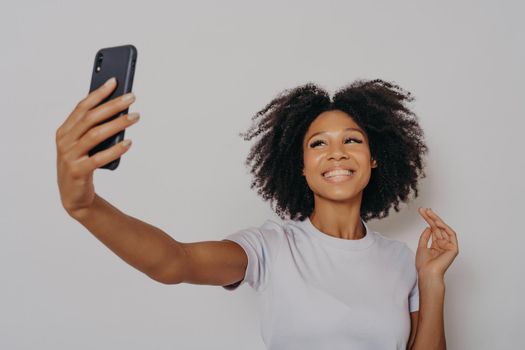 Positive cheerful dark skinned female making photo on modern smartphone camera, cheerful african woman dressed in casual white tshirt taking selfie on her mobile phone, smiling happily