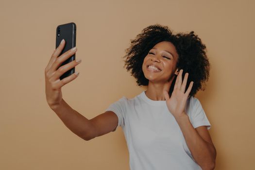 Positive happy mixed race woman waving with hand at camera during video call with friend on modern smartphone, gesturing hello to her followers in social media while posing isolated over studio wall