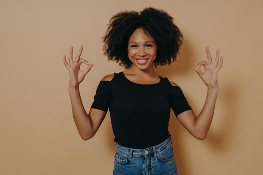 Cheerful young african female dressed in black tshirt shows ok sign okay gesture with both hands and demonstrates approval, isolated over beige background, smiling at camera and saying everything fine