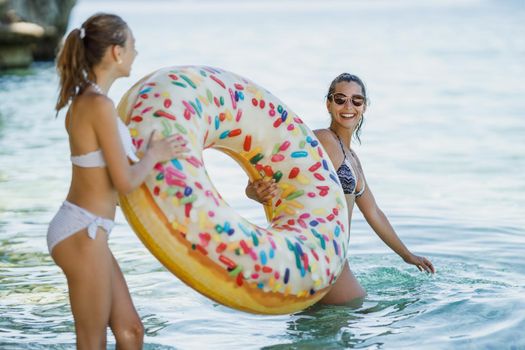 A beautiful teenage girl is enjoying with her young mother on the beach. They are having fun and bringing an inflatable rubber circle in the water to swim.