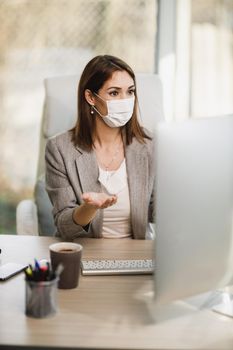An attractive young woman with surgical mask sitting alone in her office and making video call on computer. 