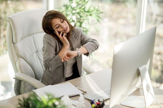 An attractive business woman looking on her wristwatch while talking on smartphone in her home office.