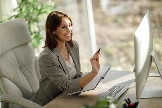 An attractive business woman checking something out of a notebook while using a computer in her office.