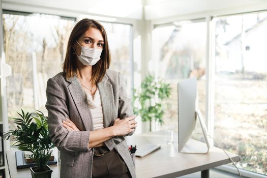 An attractive business woman with surgical mask standing in her office and looking at camera.