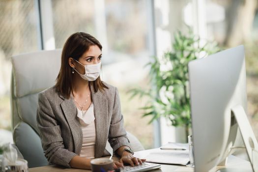 A pensive business woman with surgical mask sitting alone in her office and working on computer.
