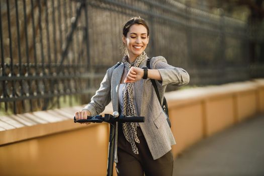 A young businesswoman with an electric push scooter going to work and looking at wristwatch.