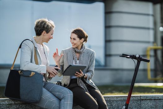 A two successful businesswomen using a digital tablet together while having a quick break in front of the office building.