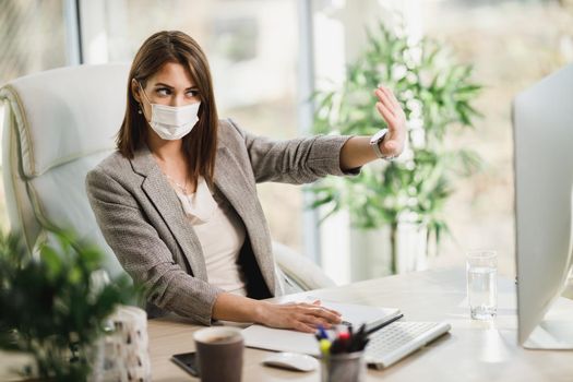 A business woman with surgical mask sitting alone in her office and working online on computer.