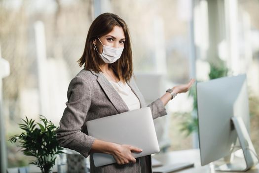 A successful young business woman with surgical mask holding laptop and standing in her office. Looking at camera.