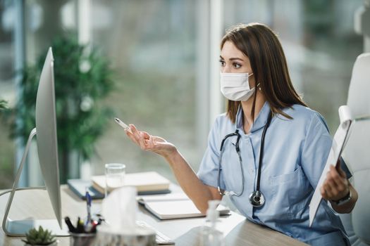 An attractive young female nurse with protective face mask working online in her consulting room during corona virus pandemic.