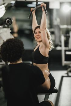 A young muscular woman is doing training at machine supporting by coach in the gym.
