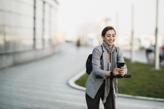 A young businesswoman using a smartphone while going to work with an electric scooter.