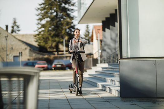 A young businesswoman looking at wristwatch and going to work with an electric scooter.