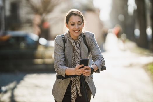 A young businesswoman using smartphone while going to work with an electric scooter.