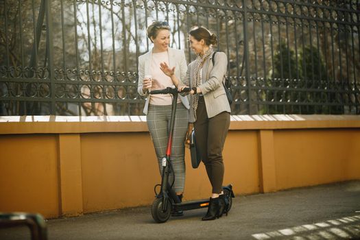 A two businesswomen with an electric scooter going to work and chatting while walking through the city.
