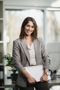 A successful young business woman holding laptop and standing in her office. Looking at camera.