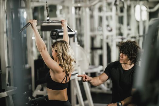 A young muscular woman is doing training at machine supporting by coach in the gym.