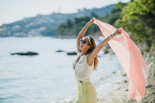 An attractive young woman is having fun and enjoying a summer vacation. She is posing with transparent scarf in her hands on the beach.