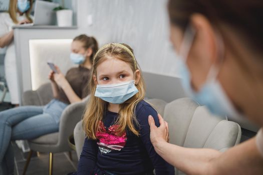 Worried little girl and her mom with face mask in waiting room at dentist's office.